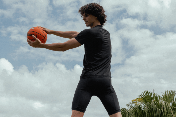 Pessoa segurando uma bola de basquete ao ar livre, vestindo camiseta e bermuda esportiva preta em tecido justo, com céu parcialmente nublado ao fundo.