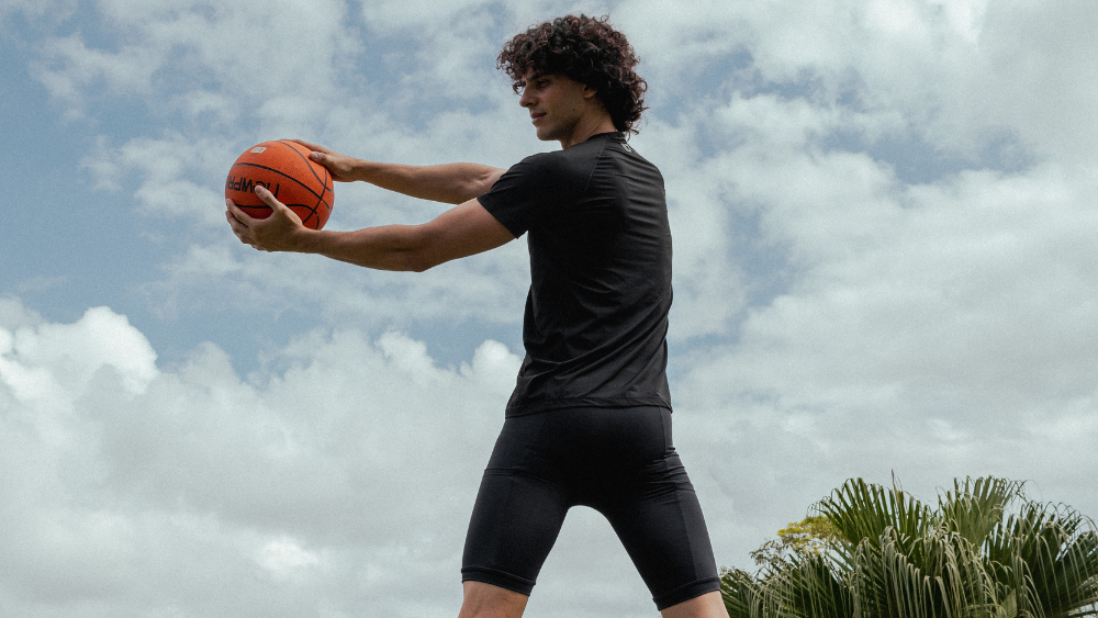 Pessoa segurando uma bola de basquete ao ar livre, vestindo camiseta e bermuda esportiva preta em tecido justo, com céu parcialmente nublado ao fundo.
