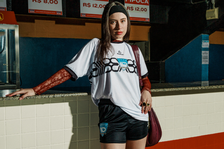 Mulher usando camisa de futebol branca e shorts pretos, em ambiente urbano, destacando o uso de tecidos tecnológicos em uniformes esportivos.