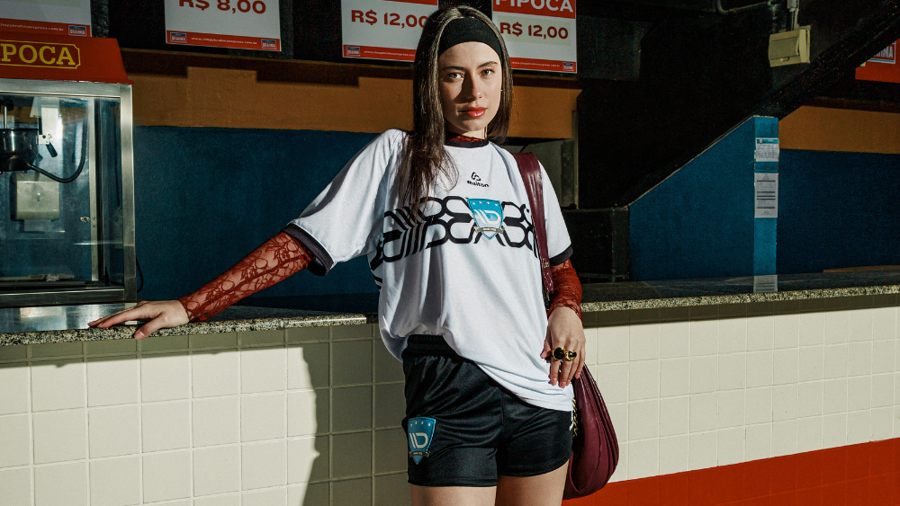 Mulher usando camisa de futebol branca e shorts pretos, em ambiente urbano, destacando o uso de tecidos tecnológicos em uniformes esportivos.