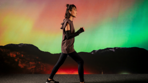 Mulher caminhando em perfil usando roupas esportivas de alta performance nas cores marrom e preto, com leggings, blusa sem mangas com capuz e manguitos. Ao fundo, uma paisagem de montanhas sob um céu com tons vibrantes de verde, laranja e rosa, remetendo à aurora boreal.