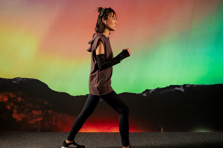 Mulher caminhando em perfil usando roupas esportivas de alta performance nas cores marrom e preto, com leggings, blusa sem mangas com capuz e manguitos. Ao fundo, uma paisagem de montanhas sob um céu com tons vibrantes de verde, laranja e rosa, remetendo à aurora boreal.
