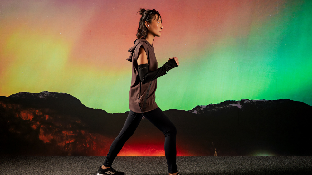 Mulher caminhando em perfil usando roupas esportivas de alta performance nas cores marrom e preto, com leggings, blusa sem mangas com capuz e manguitos. Ao fundo, uma paisagem de montanhas sob um céu com tons vibrantes de verde, laranja e rosa, remetendo à aurora boreal.