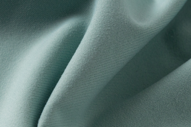 Close-up de um tecido de alta performance na cor verde suave, com superfície uniforme e toque macio, evidenciando a elasticidade, o conforto e a qualidade técnica do material utilizado na moda esportiva.